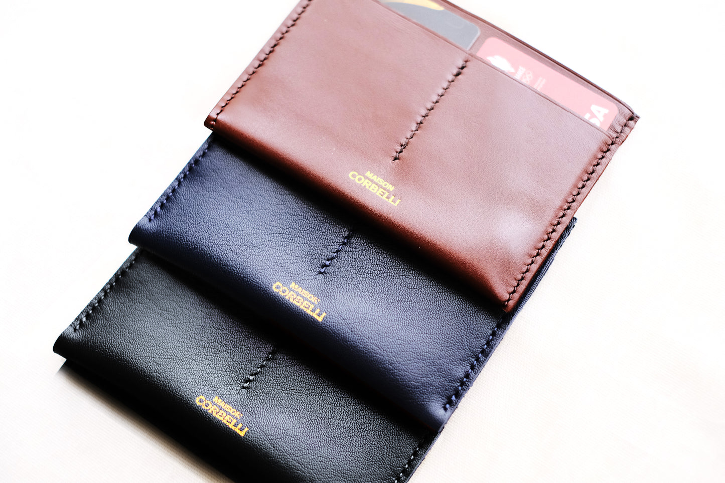 Three leather wallets, one black and one Navy blue and one cognac, with "maison Corbelli" branding in gold on a white background, Pure leather, Photo studio