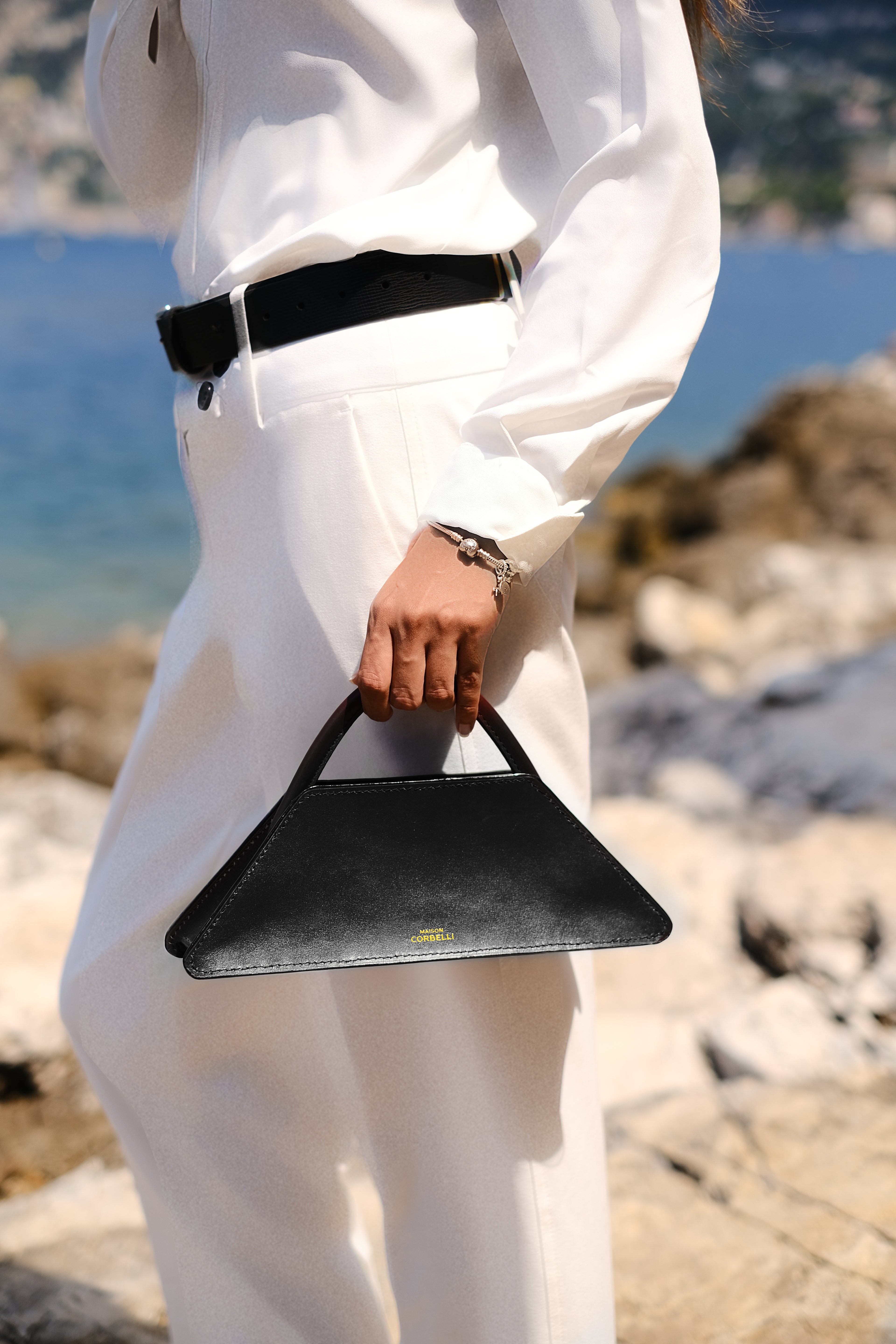 Person holding a black handbag on a rocky beach with water in the background, Woman in a white outfit standing by the sea with mountains in the background,Elie mini bag, side view, Model, street wear, Black, Clutch bag, Hand bag, mediterranean vibe, Premium leather, design, Woman model, leather, Unique design, Maison Corbelli, Luxury