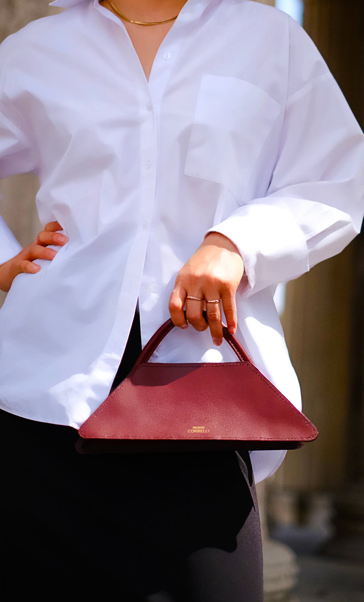 Maison Corbelli Elie handbag collection, in a burgundy colour, front view close up  woman standing in a street holding The Elie Mini clutch bag with one hand by a woman in Black and white outfit and sharp light. Golden logo with premium calf leather and a luxury look. Architectural look and Mediterranean vibes. 