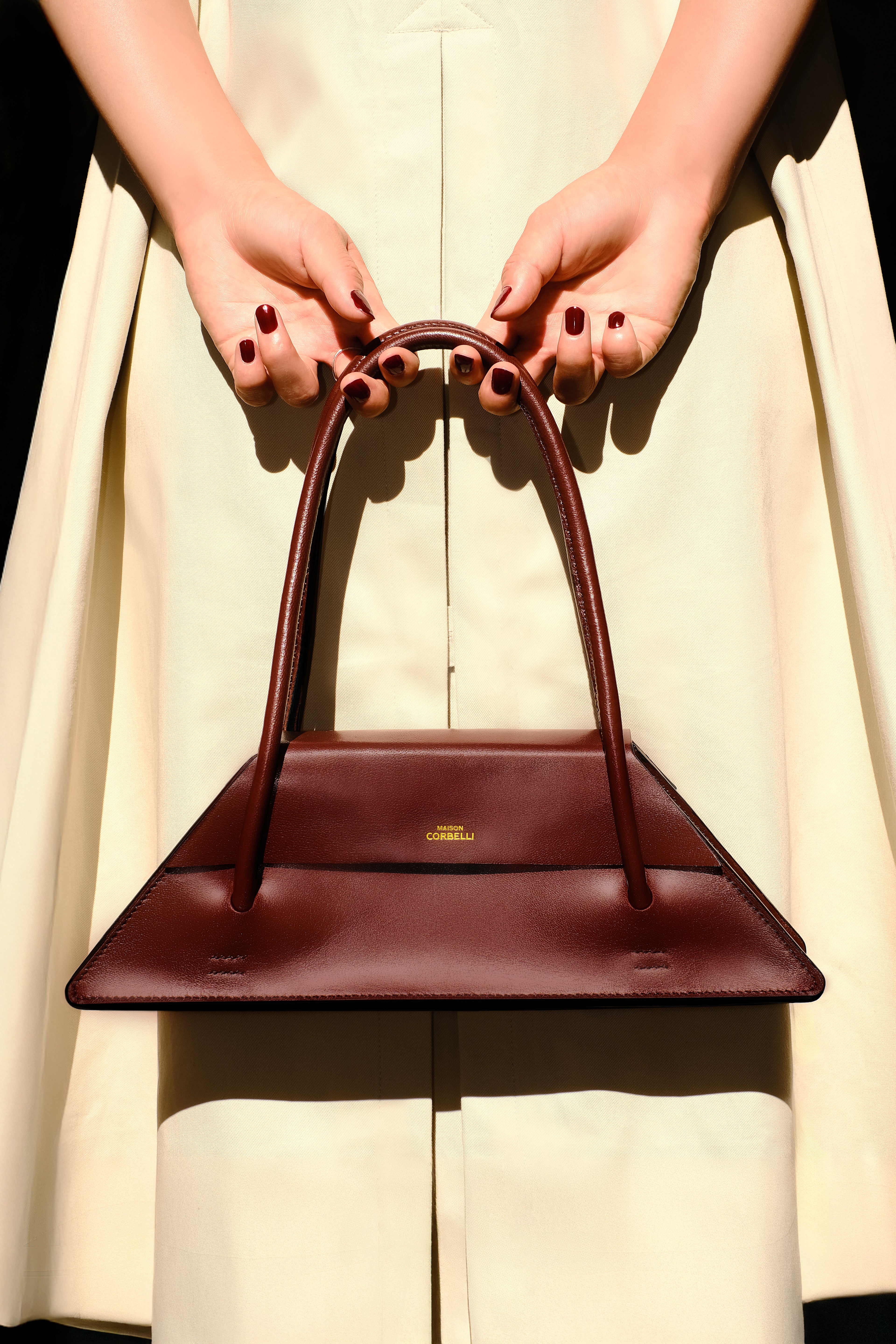 Maison Corbelli Elie handhand collection, in a burgundy colour, front view standing in a room holding with hand by a woman in a trench coat and sharp light. Golden logo with premium calf leather and a luxury look. Architectural look and mediteraneen vibes. Model carrying the Elie structured leather city bag as a shoulder bag, Original Elie Italian calf leather shoulder bag for work. Interior of Elie satchel bag showing capacity for 13-inch laptop