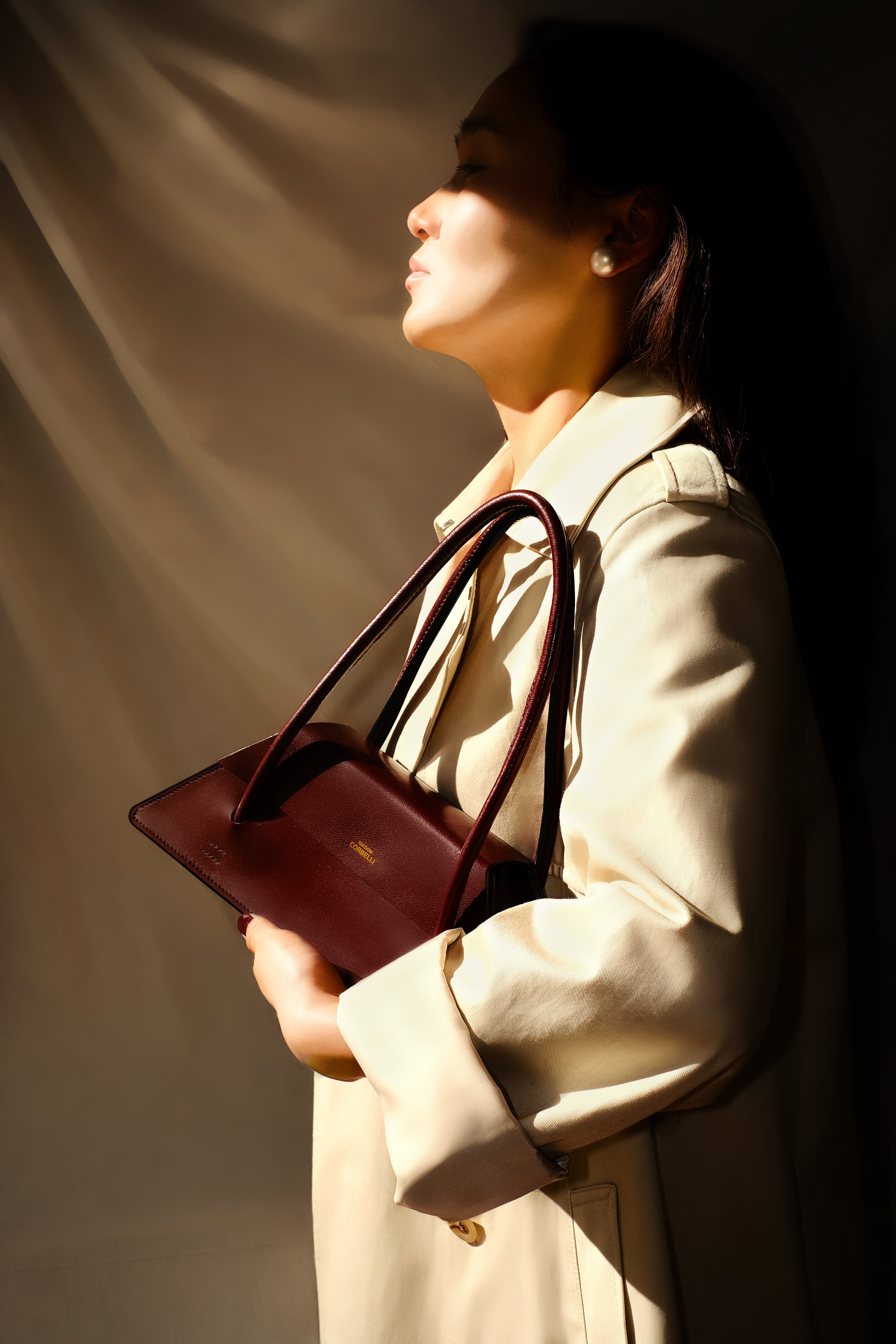 Maison Corbelli Elie handhand collection, in a burgundy colour, side view standing in a room holding with hand by a woman in a trench coat and sharp light. Golden logo with premium calf leather and a luxury look. Architectural look and mediteraneen vibes. Model carrying the Elie structured leather city bag as a shoulder bag, Original Elie Italian calf leather shoulder bag for work. Interior of Elie satchel bag showing capacity for 13-inch laptop