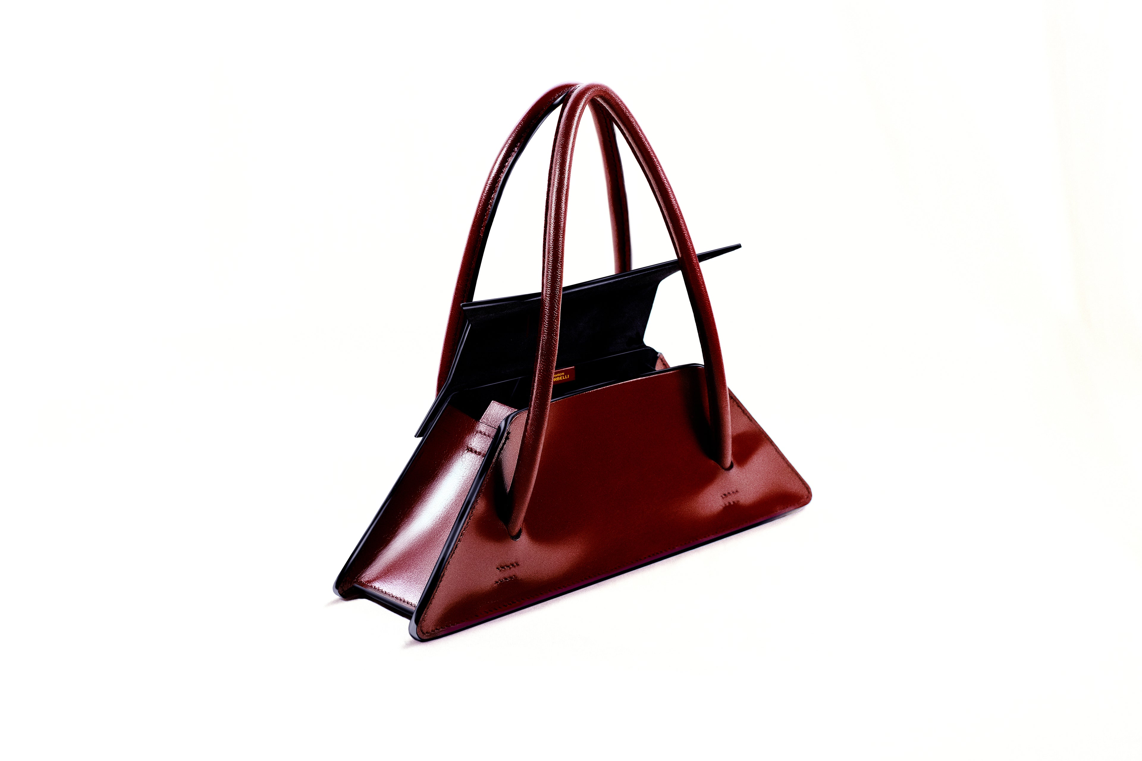 Maison Corbelli Elie handhand open collection, in a burgundy colour, side view standing in a studio with white background and sharp light. Golden logo with premium calf leather and a luxury look. Architectural look and mediteraneen vibes. Model carrying the Elie structured leather city bag as a shoulder bag, Original Elie Italian calf leather shoulder bag for work. Interior of Elie satchel bag showing capacity for 13-inch laptop