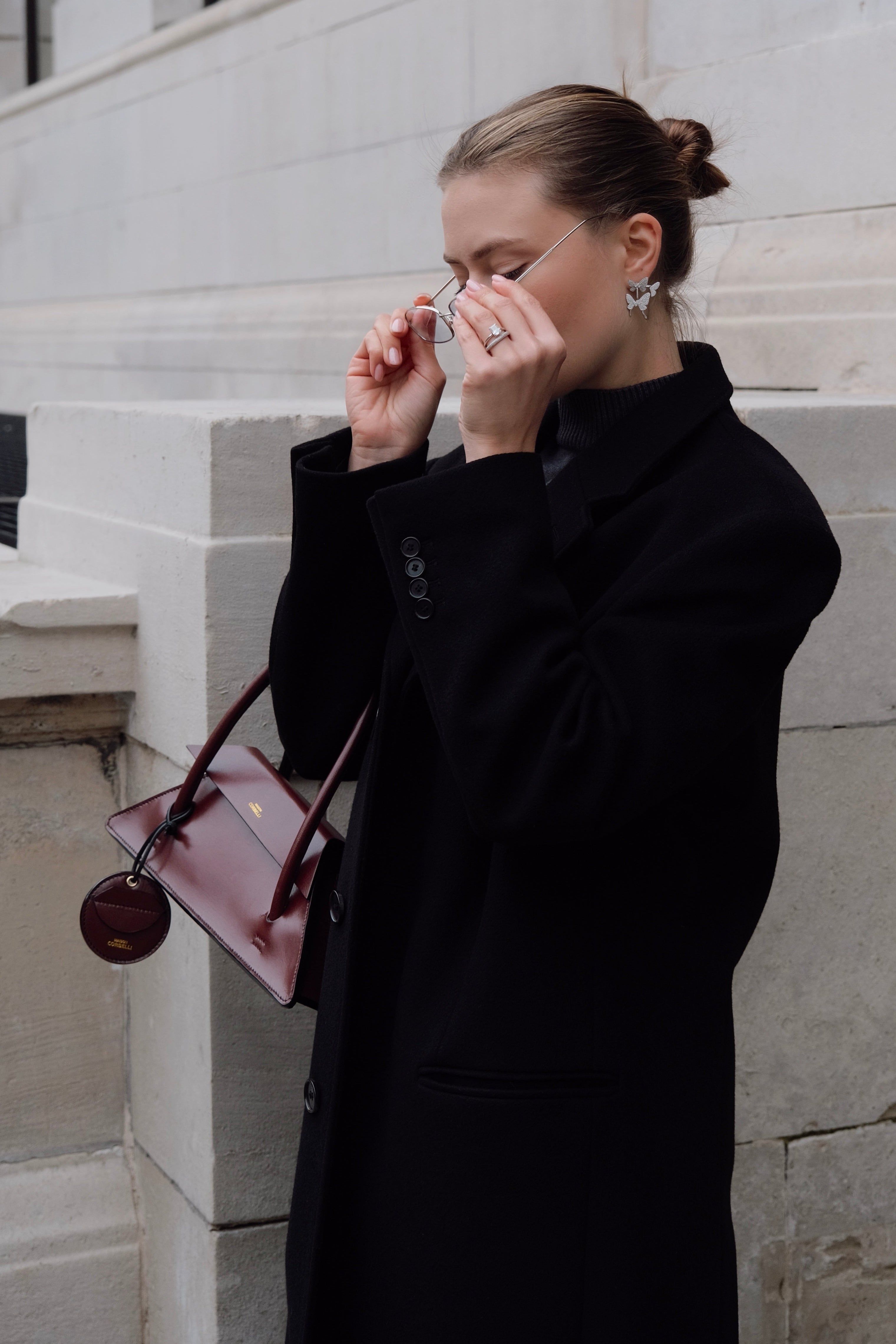 Woman in a black coat adjusting her sunglasses against a light-colored wall. Marie Latte influencer for Maison Corbelli wearing Elie bag and the Oto coin holder