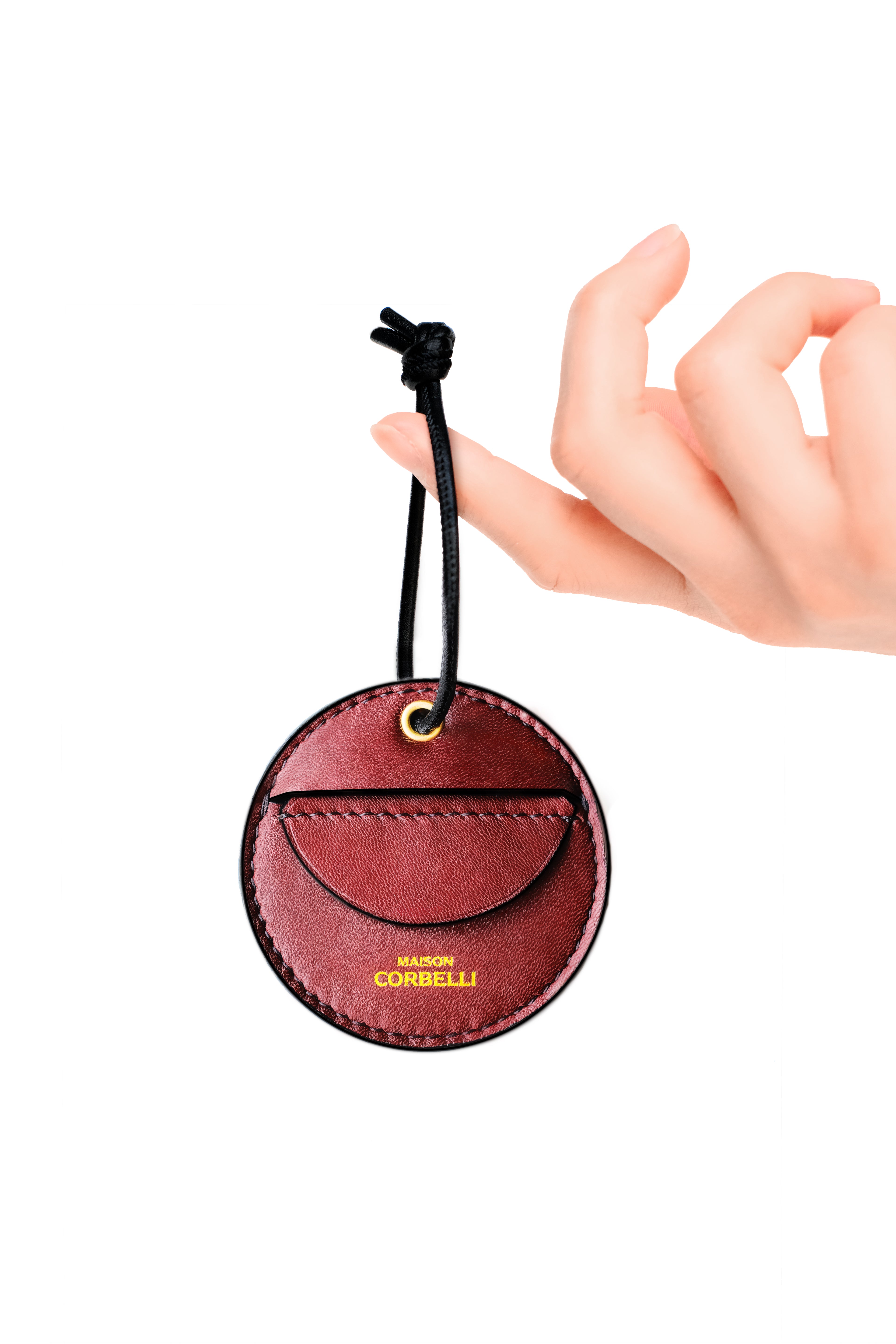 Maison Corbelli, Round leather coin holder with a hand holding it on a white background, Oto, coin holder, Burgundy, red, gift for christmas, present, gold-colored accessories, coin purse, a finger holds it, Front view, Model, Studio, Burgundy, bag, mediterranean vibe, Premium leather, Unique design