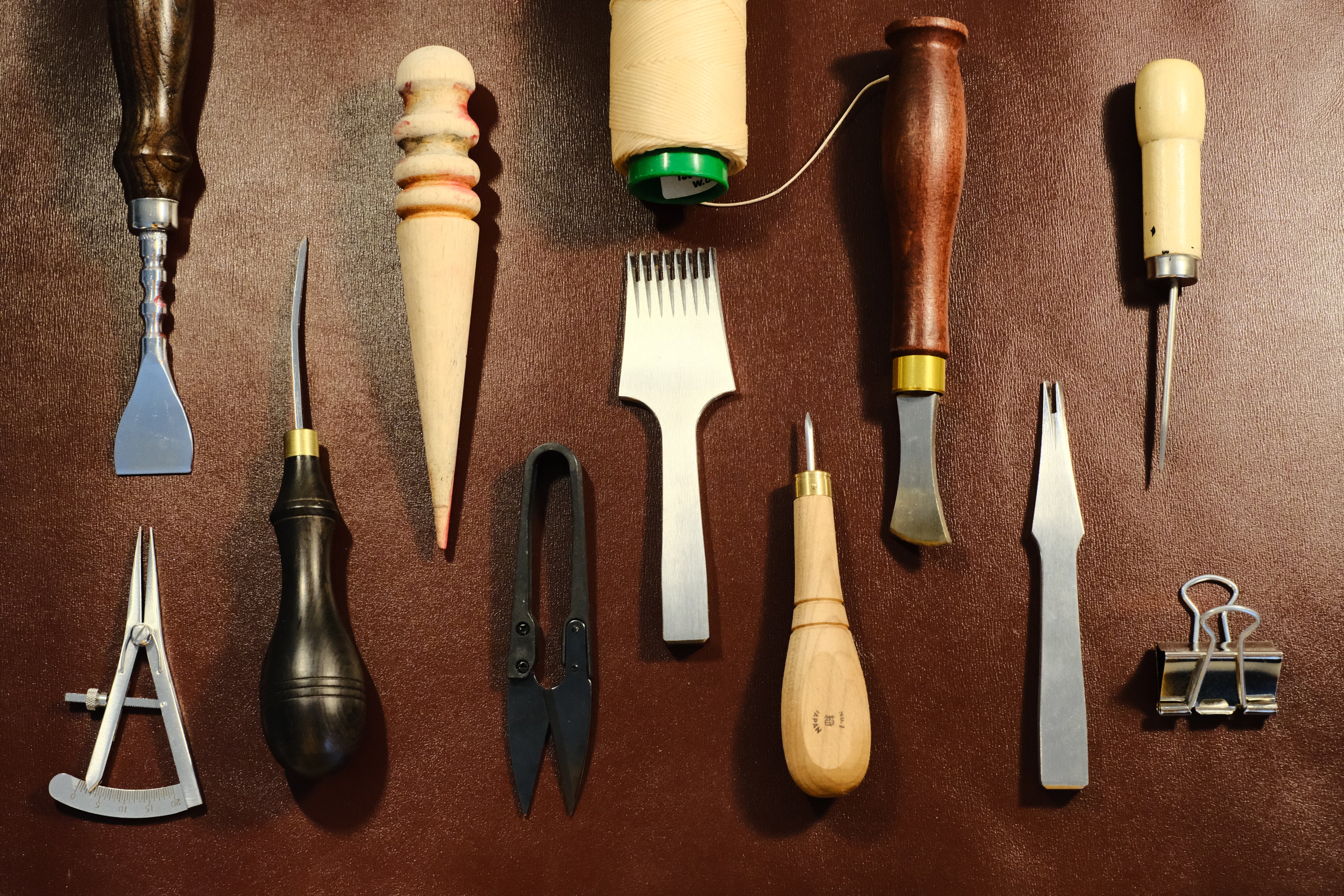 Top view of luxury leathercraft tools on a leather background chisel, thread, and awl arranged beside a piece of leather showcasing precise saddle stitching and artisan craftsmanship. Maison Corbelli, 
brown background. Sac à main et outils