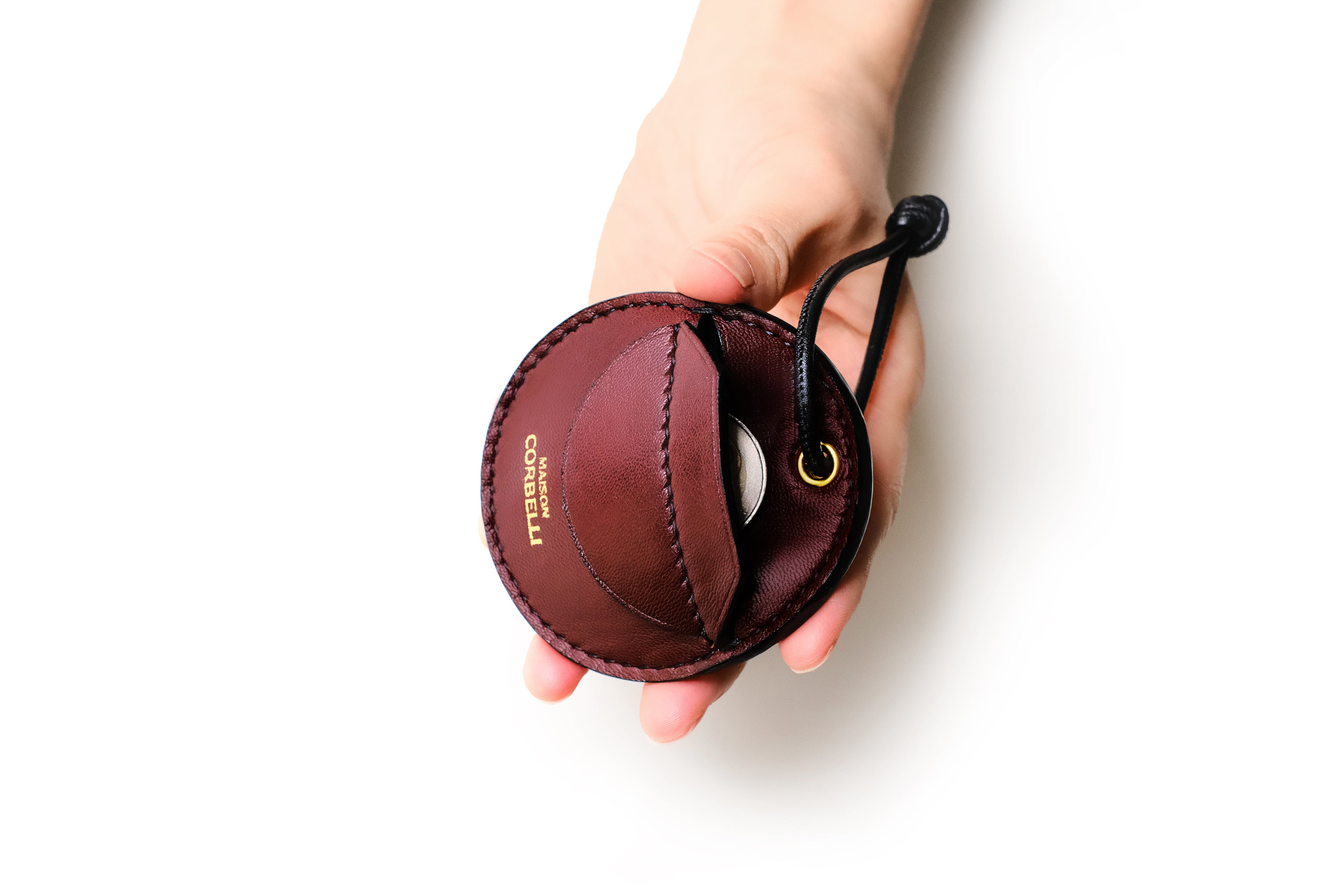 Blue leather luggage tag with 'MAISON CORBELLI' branding on a white background, Round leather coin holder with a hand holding it on a white background, Oto, coin holder, Burgundy, red, dark red, gift for christmas, present, gold-colored accessories, coin purse, a finger holds it, Front view, Model, Studio, Burgundy, bag, mediterranean vibe, Premium leather, Unique design